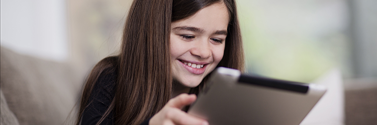 A young girl sitting on a sofa, smiling towards a tablet that is in her hands