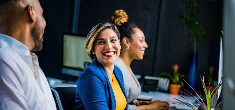 Three office workers with hands on laptops, one female colleague smiling towards the camera and a colleague