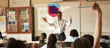 Teacher standing at the front of a classroom with students raising hands.