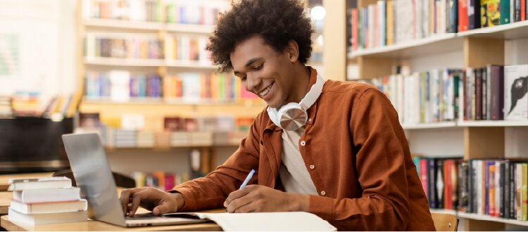 Person studying at a laptop in a library, writing notes with bookshelves in the background.