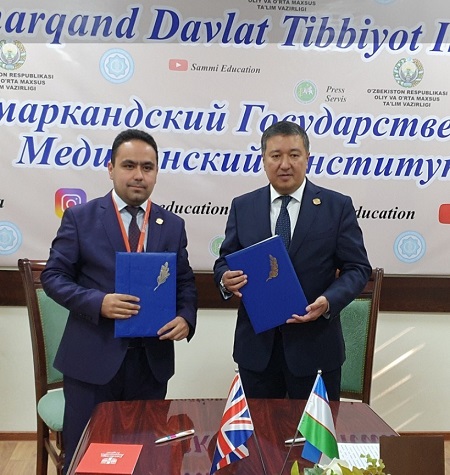 Two gentlemen in formal jackets standing in front of a sponsored wall and behind a table with a union jack flag and Uzbekistan flag on the surface. Both gentlemen are holding a blue book each