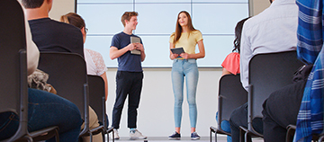 Dos alumnos hacen una presentación frente a una clase.